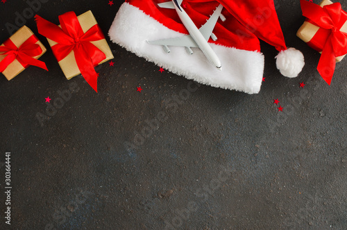 Christmas or New Year travel concept. Toy airplane on Santa hat and gift boxes on dark concrete.