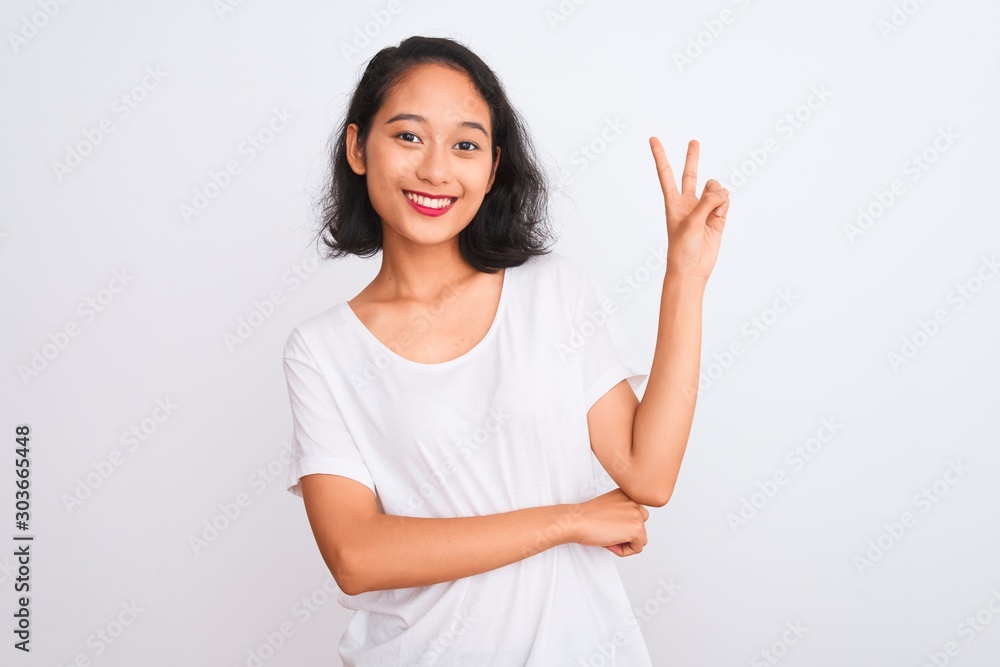 Young chinese woman wearing casual t-shirt standing over isolated white background smiling with happy face winking at the camera doing victory sign. Number two. - obrazy, fototapety, plakaty 