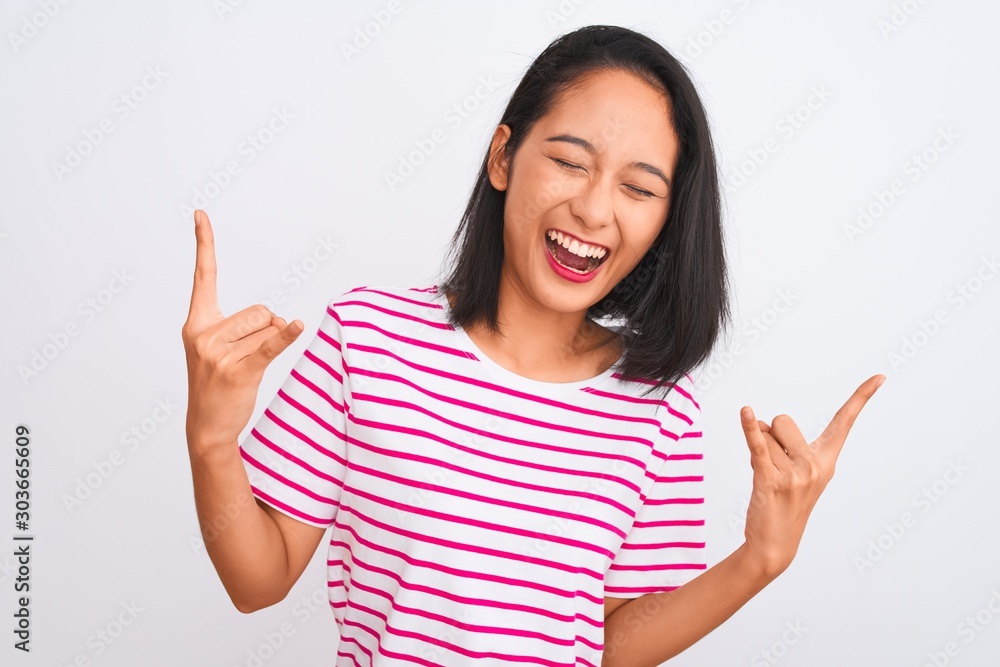 Young chinese woman wearing striped t-shirt standing over isolated white background shouting with crazy expression doing rock symbol with hands up. Music star. Heavy concept. Young chinese woman wearing striped t-shirt standing over isolated white background shouting with crazy expression doing rock symbol with hands up. Music star. Heavy concept.