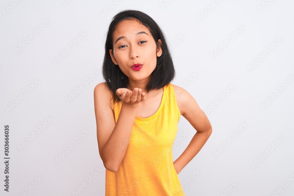 Young chinese woman wearing yellow casual t-shirt standing over isolated white background looking at the camera blowing a kiss with hand on air being lovely and sexy. Love expression. Young chinese woman wearing yellow casual t-shirt standing over isolated white background looking at the camera blowing a kiss with hand on air being lovely and sexy. Love expression.