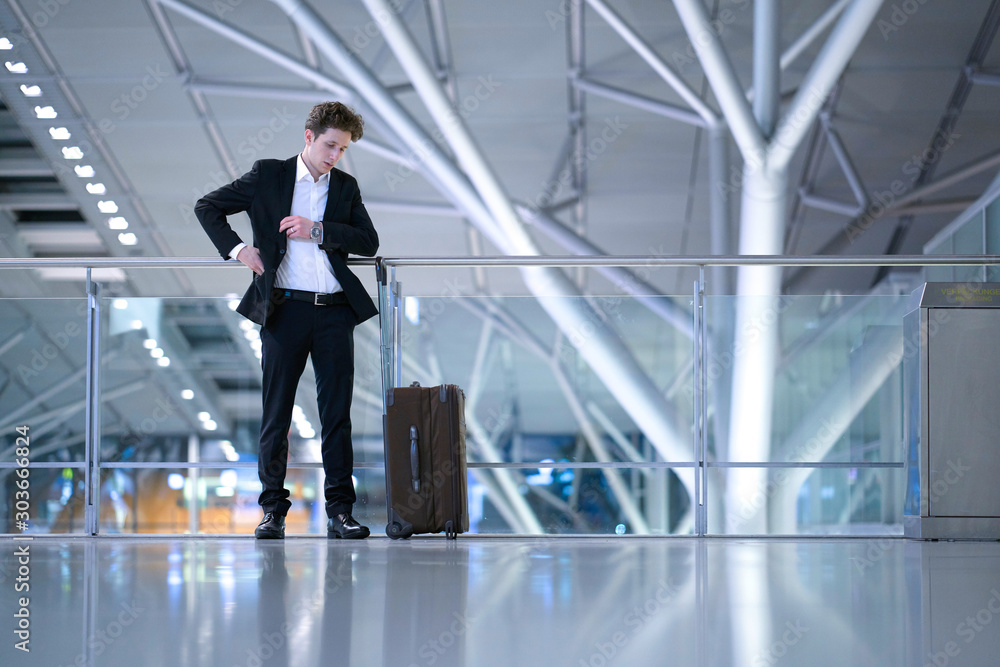 Young businessman standing inside the airport in front of a glass guard railing with his rolling case, searching something in his suit pocket Young businessman standing inside the airport in front of a glass guard railing with his rolling case, searching something in his suit pocket