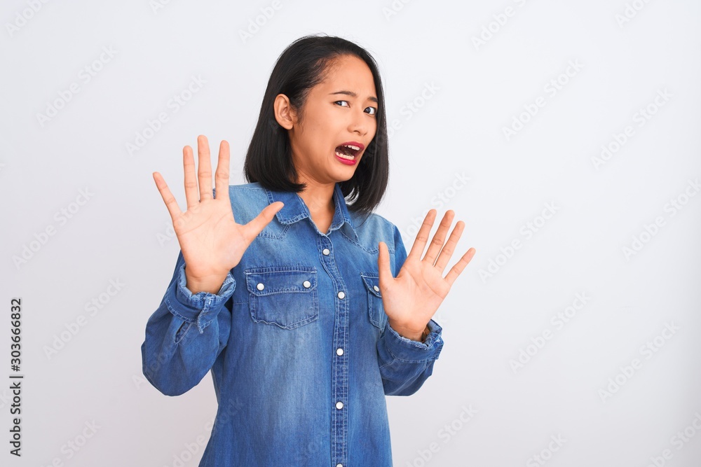 Young beautiful chinese woman wearing denim shirt standing over isolated white background afraid and terrified with fear expression stop gesture with hands, shouting in shock. Panic concept. Young beautiful chinese woman wearing denim shirt standing over isolated white background afraid and terrified with fear expression stop gesture with hands, shouting in shock. Panic concept.