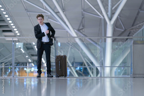 Young businessman standing inside the airport in front of a glass guard railing with his rolling case, searching something in his suit pocket