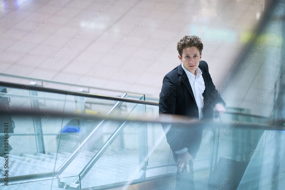 Young businessman on an escalator in the airport, one hand at his rolling case Young businessman on an escalator in the airport, one hand at his rolling case
