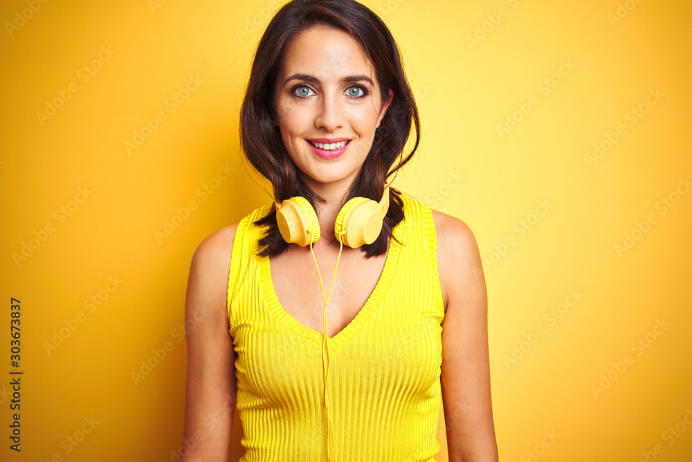 Young beautiful woman wearing headphones over yellow isolated background with a happy and cool smile on face. Lucky person. Young beautiful woman wearing headphones over yellow isolated background with a happy and cool smile on face. Lucky person.
