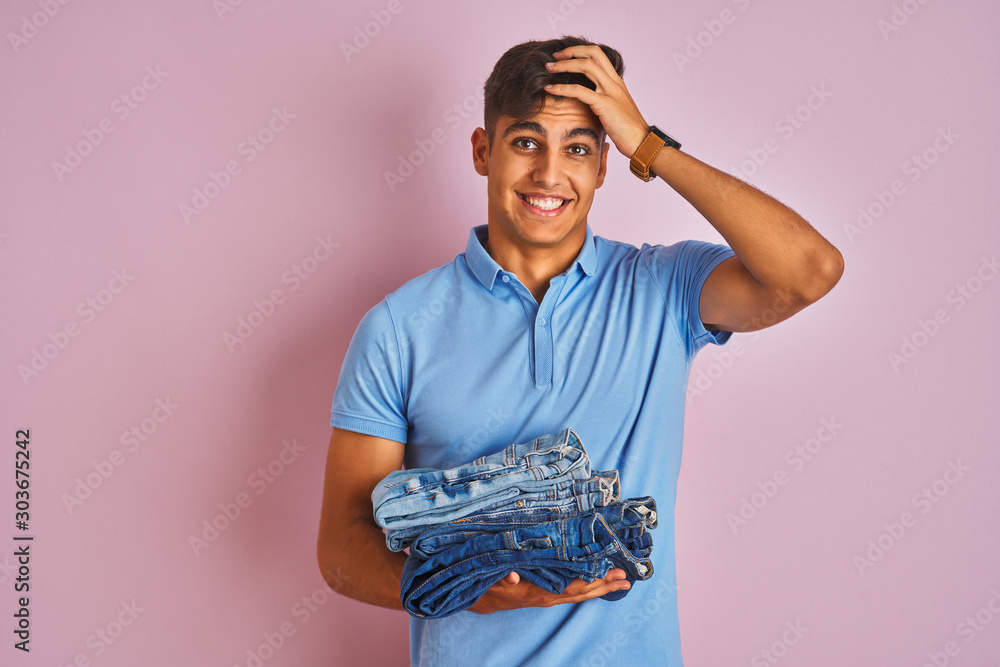 Φωτογραφία Young indian shopkeeper man holding folded jeans standing over isolated pink background stressed with hand on head, shocked with shame and surprise face, angry and frustrated