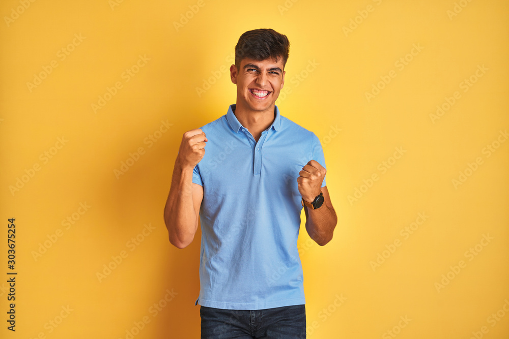 Young indian man wearing blue polo standing over isolated yellow background very happy and excited doing winner gesture with arms raised, smiling and screaming for success. Celebration concept. Young indian man wearing blue polo standing over isolated yellow background very happy and excited doing winner gesture with arms raised, smiling and screaming for success. Celebration concept.
