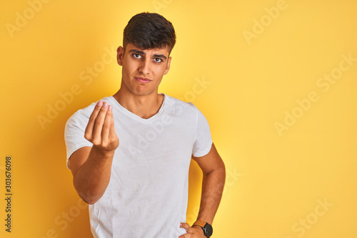 Young indian man wearing white t-shirt standing over isolated yellow background Doing Italian gesture with hand and fingers confident expression