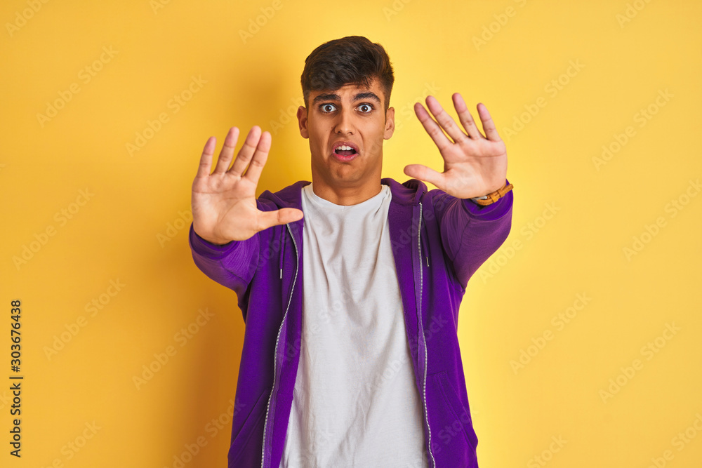 Young indian man wearing purple sweatshirt standing over isolated yellow background afraid and terrified with fear expression stop gesture with hands, shouting in shock. Panic concept. Young indian man wearing purple sweatshirt standing over isolated yellow background afraid and terrified with fear expression stop gesture with hands, shouting in shock. Panic concept.