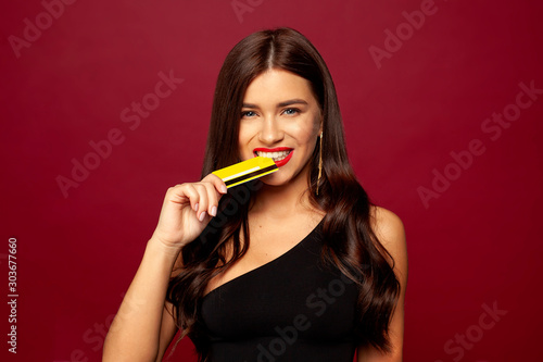  beautiful brunette model is standing on a red background in a small black dress, with red lipstick and smiling, she is holding a credit card in her teeth, Merry Christmas and Happy New Year