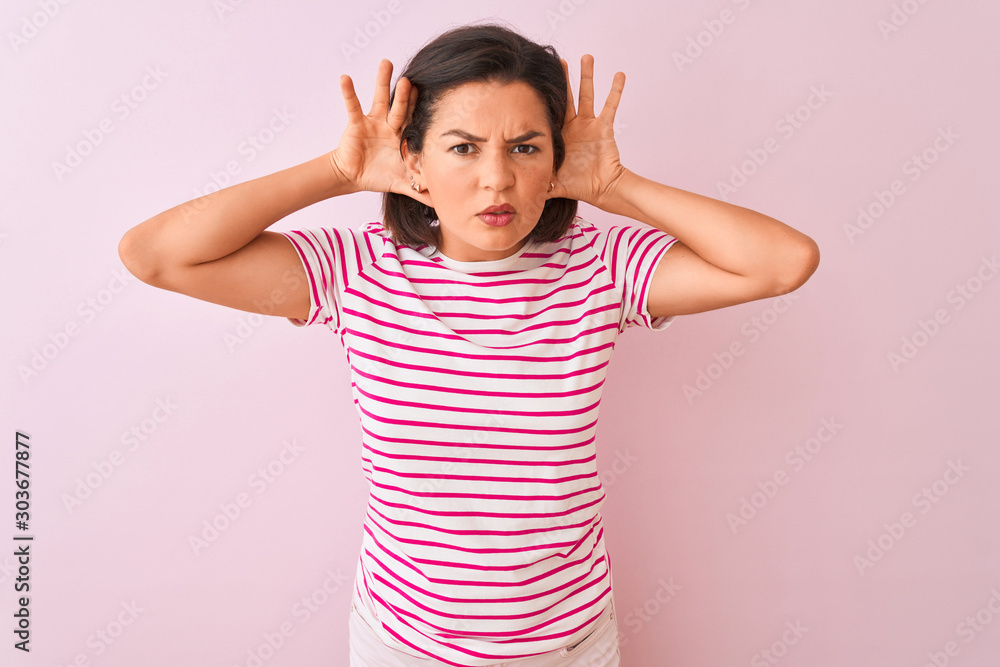 Young beautiful woman wearing striped t-shirt standing over isolated pink background Trying to hear both hands on ear gesture, curious for gossip. Hearing problem, deaf Young beautiful woman wearing striped t-shirt standing over isolated pink background Trying to hear both hands on ear gesture, curious for gossip. Hearing problem, deaf