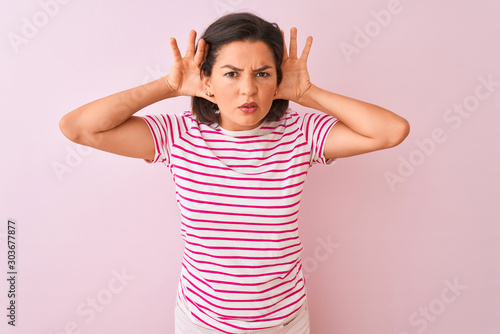 Young beautiful woman wearing striped t-shirt standing over isolated pink background Trying to hear both hands on ear gesture, curious for gossip. Hearing problem, deaf