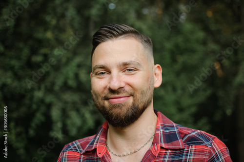 Close up portrait of young bearded man in red shirt looking at the camera with smile in the park on trees background