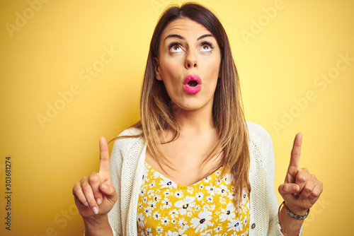 Young beautiful woman wearing jacket standing over yellow isolated background amazed and surprised looking up and pointing with fingers and raised arms.