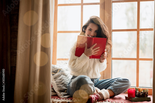 brunette woman in warm sweater and socks reading book sitting on windowsill in room decorated for celebrating new year and christmas looking happy festive mood