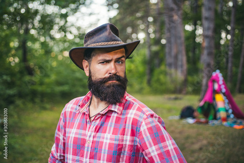 Guy in forest. Handsome bearded male. Natural background. Brutal brunette bearded man in hat on a background of trees. Mature hipster with beard. brutal man with beard. © Volodymyr