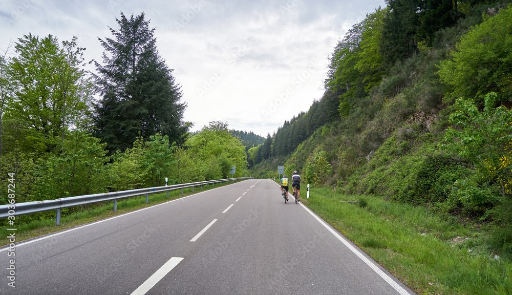 A highway through a forest with turns and two cyclist riders ride along it. Schwarzwald forest road and cyclists. Germany A highway through a forest with turns and two cyclist riders ride along it. Schwarzwald forest road and cyclists. Germany