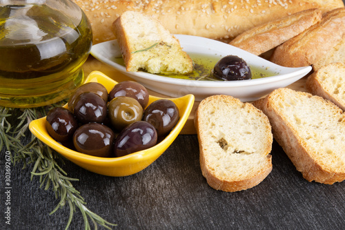 Close up of yellow bowl with olives and rosemary. Olive oil and bread on wooden background. Greek black olives