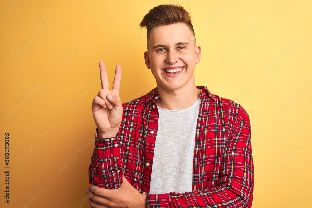 Young handsome man wearing casual shirt standing over isolated yellow background smiling with happy face winking at the camera doing victory sign. Number two. Young handsome man wearing casual shirt standing over isolated yellow background smiling with happy face winking at the camera doing victory sign. Number two.