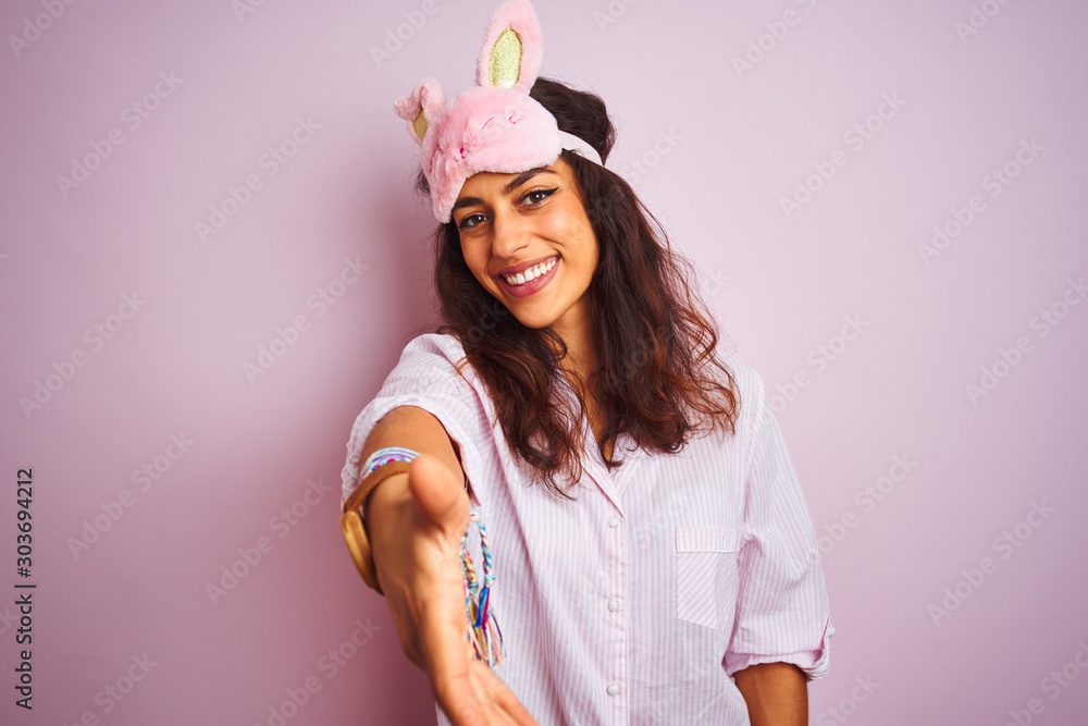 Young beautiful woman wearing pajama and sleep mask over isolated pink background smiling friendly offering handshake as greeting and welcoming. Successful business.