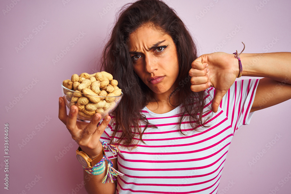 Young beautiful woman holding bowl with peanuts over isolated pink background with angry face, negative sign showing dislike with thumbs down, rejection concept Young beautiful woman holding bowl with peanuts over isolated pink background with angry face, negative sign showing dislike with thumbs down, rejection concept