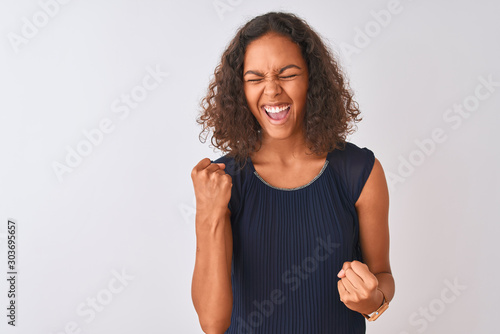 Young brazilian woman wearing blue dress standing over isolated white background very happy and excited doing winner gesture with arms raised, smiling and screaming for success. Celebration concept.