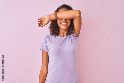 Young brazilian woman wearing t-shirt standing over isolated pink background covering eyes with arm smiling cheerful and funny. Blind concept.