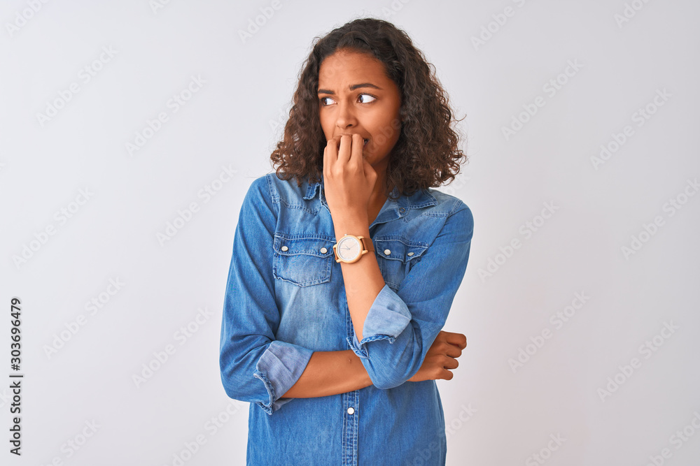 Young brazilian woman wearing denim shirt standing over isolated white background looking stressed and nervous with hands on mouth biting nails. Anxiety problem. Young brazilian woman wearing denim shirt standing over isolated white background looking stressed and nervous with hands on mouth biting nails. Anxiety problem.