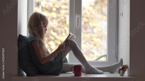 Child girl in turquoise dress reading book on windowsill with teddy bear