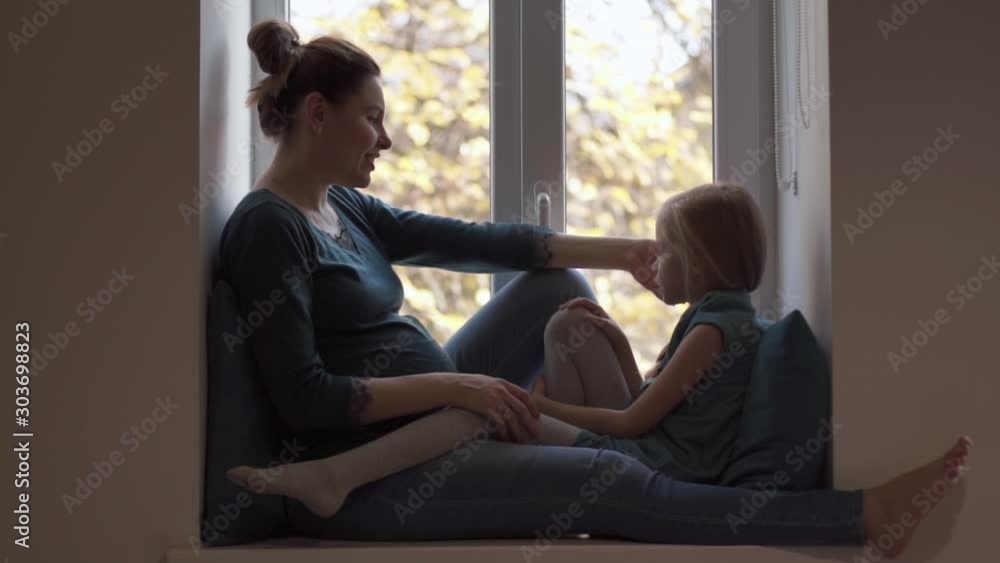 Pregnant woman caress daughter in turquoise clothes on windowsill Pregnant woman caress daughter in turquoise clothes on windowsill