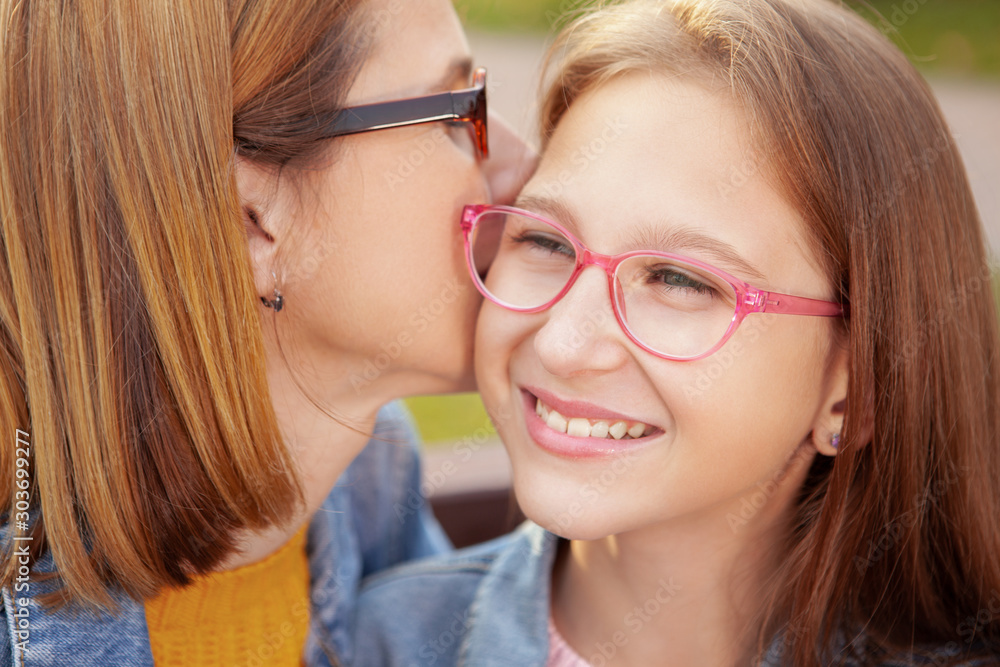 Close up of a lovely young girl smiling joyfully as her mother kissing her on the cheek. Mom and daughter cuddling outdoors Close up of a lovely young girl smiling joyfully as her mother kissing her on the cheek. Mom and daughter cuddling outdoors