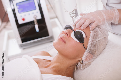 Close up of a beautiful woman in protective eyeglasses getting facial laser treatment by professional cosmetologist 