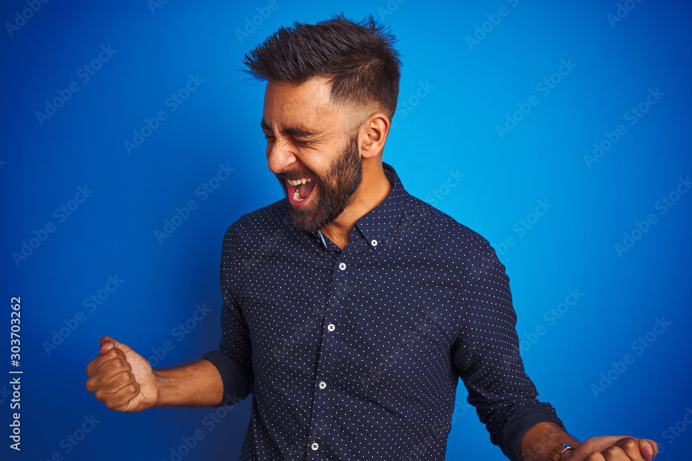 Young indian elegant man wearing shirt standing over isolated blue background very happy and excited doing winner gesture with arms raised, smiling and screaming for success. Celebration concept.