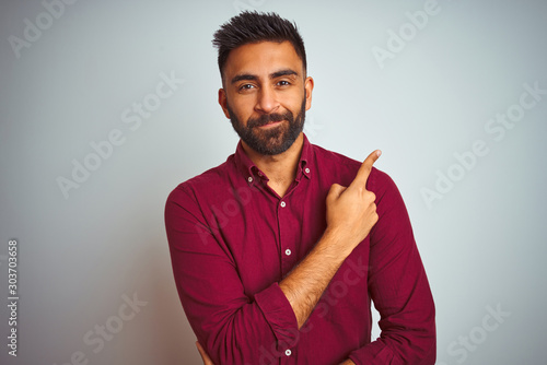 Young indian man wearing red elegant shirt standing over isolated grey background Pointing with hand finger to the side showing advertisement, serious and calm face