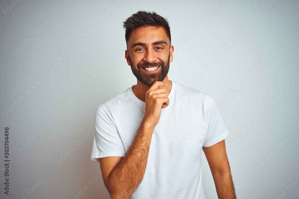 Young indian man wearing t-shirt standing over isolated white background with hand on chin thinking about question, pensive expression. Smiling and thoughtful face. Doubt concept. Young indian man wearing t-shirt standing over isolated white background with hand on chin thinking about question, pensive expression. Smiling and thoughtful face. Doubt concept.