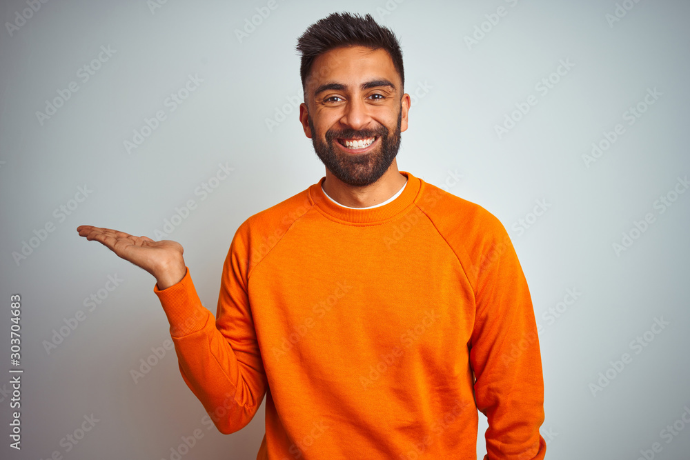 Φωτογραφία Young indian man wearing orange sweater over isolated white background smiling cheerful presenting and pointing with palm of hand looking at the camera