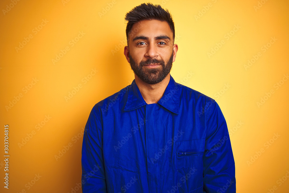 Handsome indian worker man wearing uniform over isolated yellow background with serious expression on face. Simple and natural looking at the camera. Handsome indian worker man wearing uniform over isolated yellow background with serious expression on face. Simple and natural looking at the camera.