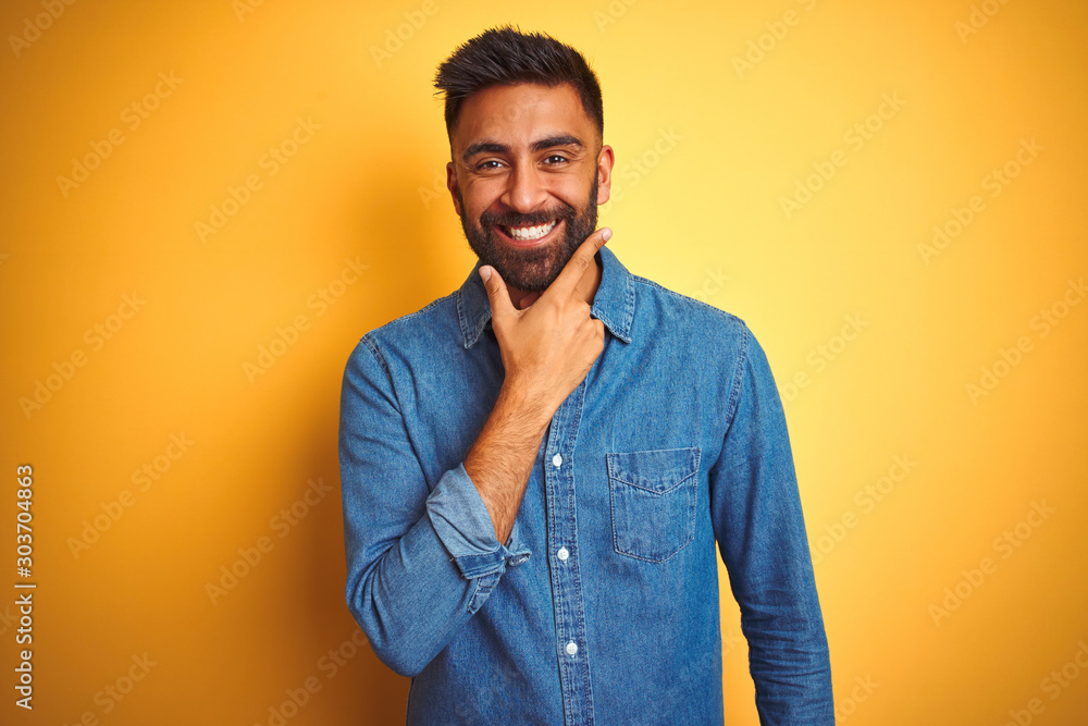 Young indian man wearing denim shirt standing over isolated yellow background looking confident at the camera smiling with crossed arms and hand raised on chin. Thinking positive. Young indian man wearing denim shirt standing over isolated yellow background looking confident at the camera smiling with crossed arms and hand raised on chin. Thinking positive.