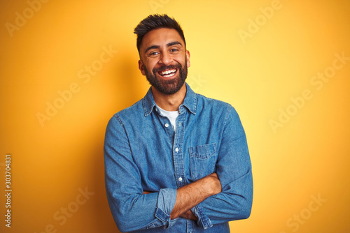 Young indian man wearing denim shirt standing over isolated yellow background happy face smiling with crossed arms looking at the camera. Positive person.