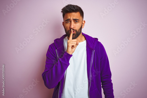 Young indian man wearing purple sweatshirt standing over isolated pink background asking to be quiet with finger on lips. Silence and secret concept.