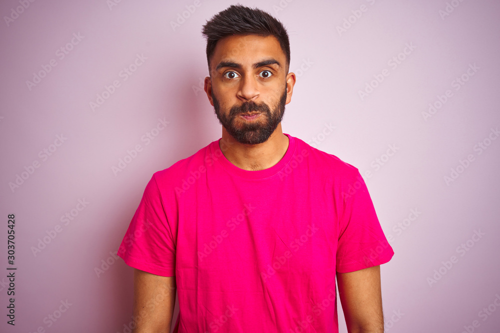 Young indian man wearing t-shirt standing over isolated pink background puffing cheeks with funny face. Mouth inflated with air, crazy expression. Young indian man wearing t-shirt standing over isolated pink background puffing cheeks with funny face. Mouth inflated with air, crazy expression.