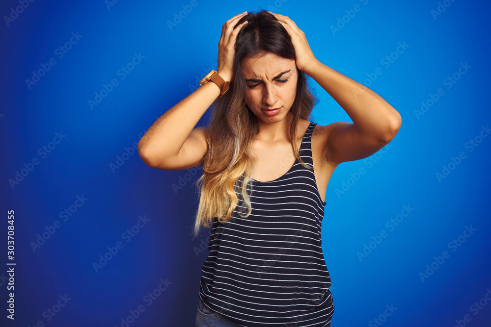 Young beautiful woman wearing stripes t-shirt over blue isolated background suffering from headache desperate and stressed because pain and migraine. Hands on head. Young beautiful woman wearing stripes t-shirt over blue isolated background suffering from headache desperate and stressed because pain and migraine. Hands on head.