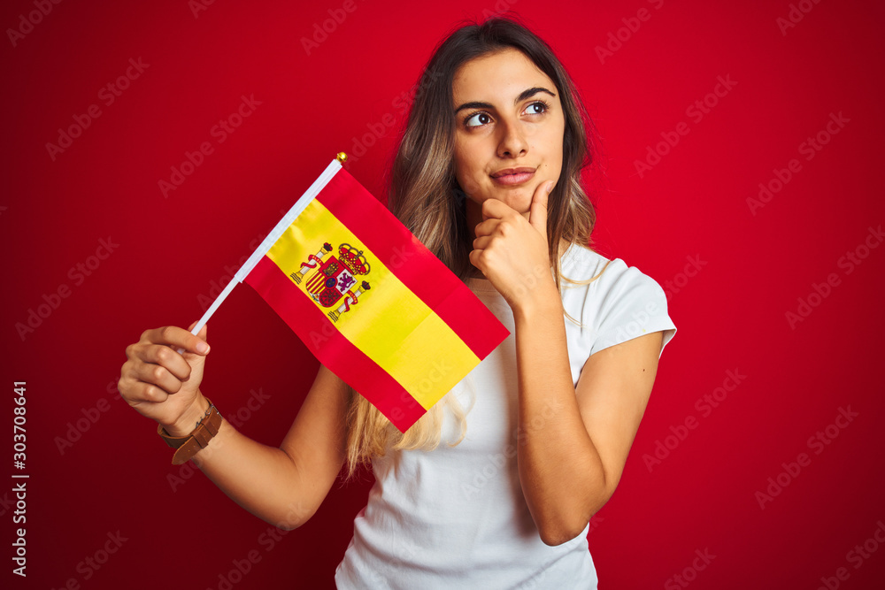Young beautiful woman holding spanish flag over red isolated background serious face thinking about question, very confused idea Young beautiful woman holding spanish flag over red isolated background serious face thinking about question, very confused idea