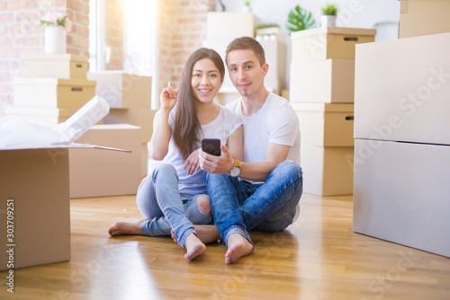 Beautiful couple sitting on the floor using smartphone at new home around cardboard boxes surprised with an idea or question pointing finger with happy face, number one © Krakenimages.com