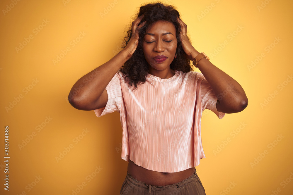 African american woman wearing pink t-shirt standing over isolated yellow background suffering from headache desperate and stressed because pain and migraine. Hands on head. African american woman wearing pink t-shirt standing over isolated yellow background suffering from headache desperate and stressed because pain and migraine. Hands on head.