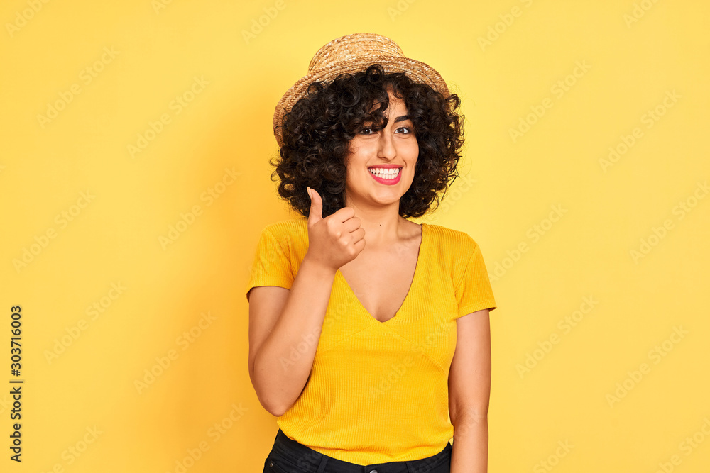 Young arab woman with curly hair wearing t-shirt and hat over isolated yellow background doing happy thumbs up gesture with hand. Approving expression looking at the camera with showing success. Young arab woman with curly hair wearing t-shirt and hat over isolated yellow background doing happy thumbs up gesture with hand. Approving expression looking at the camera with showing success.