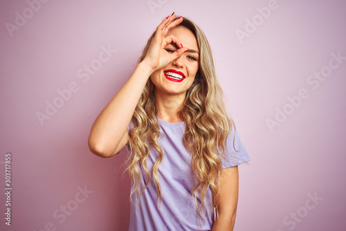 Young beautiful woman wearing purple t-shirt standing over pink isolated background doing ok gesture with hand smiling, eye looking through fingers with happy face.