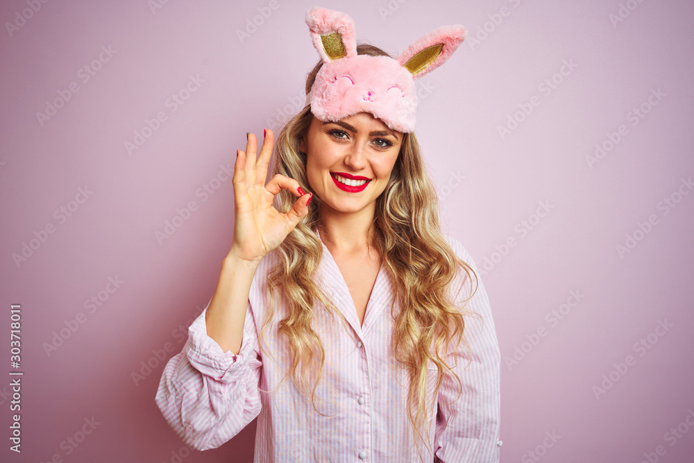 Young beautiful woman wearing pajama and sleep mask over pink isolated background smiling positive doing ok sign with hand and fingers. Successful expression.