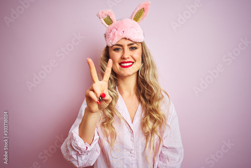 Young beautiful woman wearing pajama and sleep mask over pink isolated background showing and pointing up with fingers number two while smiling confident and happy.