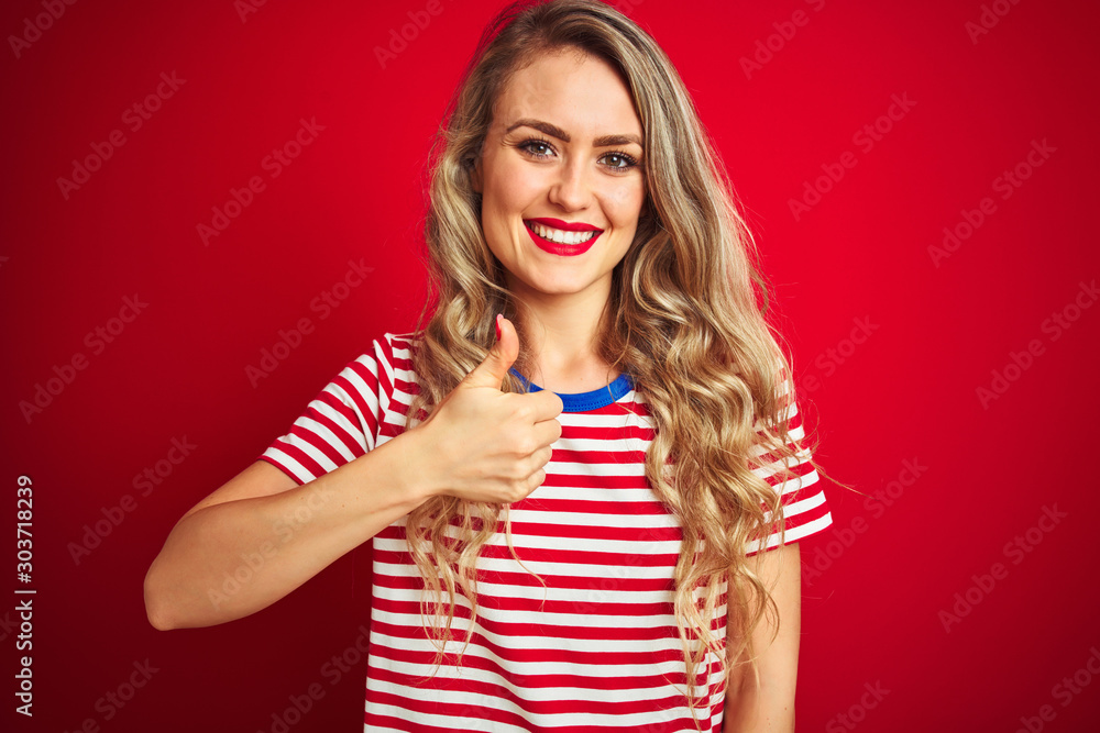 Young beautiful woman wearing stripes t-shirt standing over red isolated background doing happy thumbs up gesture with hand. Approving expression looking at the camera with showing success. Young beautiful woman wearing stripes t-shirt standing over red isolated background doing happy thumbs up gesture with hand. Approving expression looking at the camera with showing success.